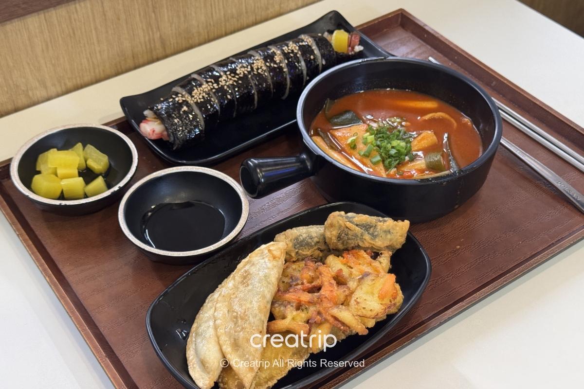 在釜山金海机场餐厅桌上的韩式套餐：紫菜饭卷、辣炒年糕汤、综合炸物与小菜，搭配酱料与餐具，呈现登机前可快速品尝的道地机场美食场景。