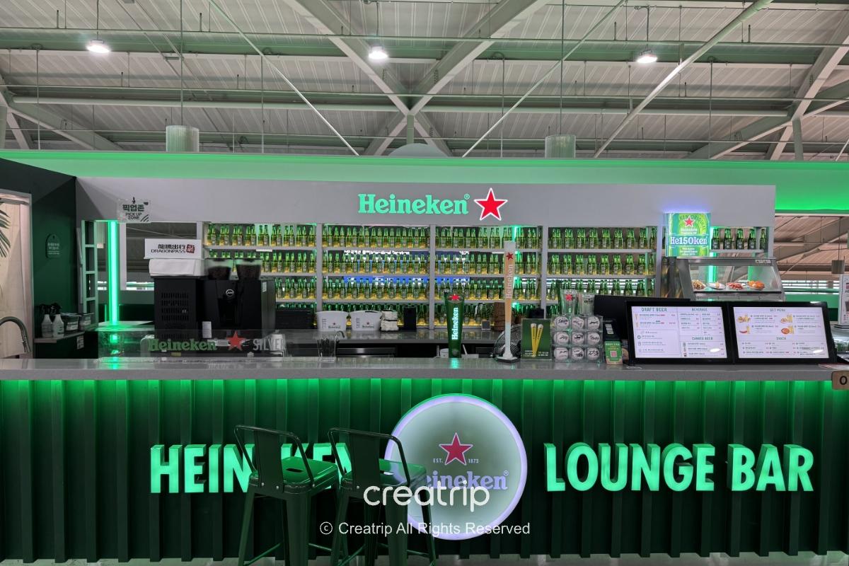 釜山金海机场内的海尼根Lounge Bar吧台，绿色霓虹灯与酒瓶墙营造酒吧氛围，吧台上有点餐萤幕与座位，适合登机前放松小酌或享用炸物。