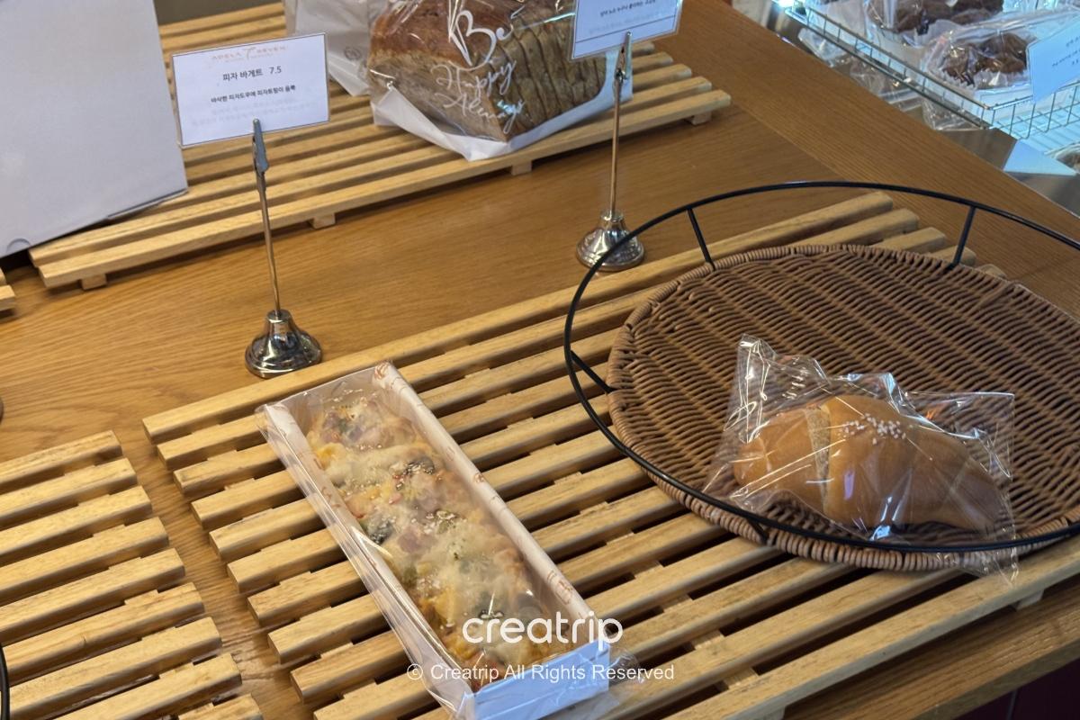 ADELA 7面包店柜台展示包装面包与长条披萨，木质展示架与藤篮强调手作感，右下有Creatrip浮水印。