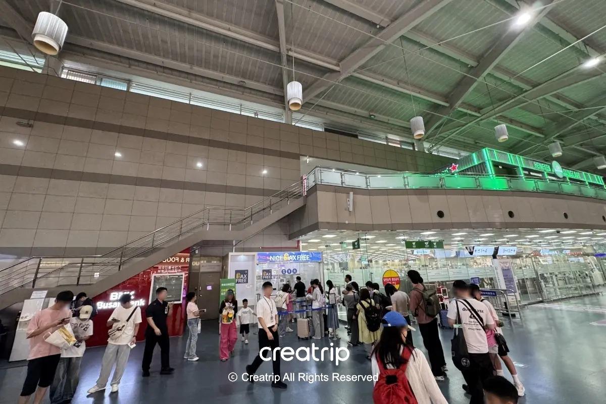 釜山金海機場出發大廳一景：旅客排隊辦理手續與候位，樓上可見綠色霓虹的酒吧招牌與樓梯通往餐飲區，示意機場內餐廳分布與動線