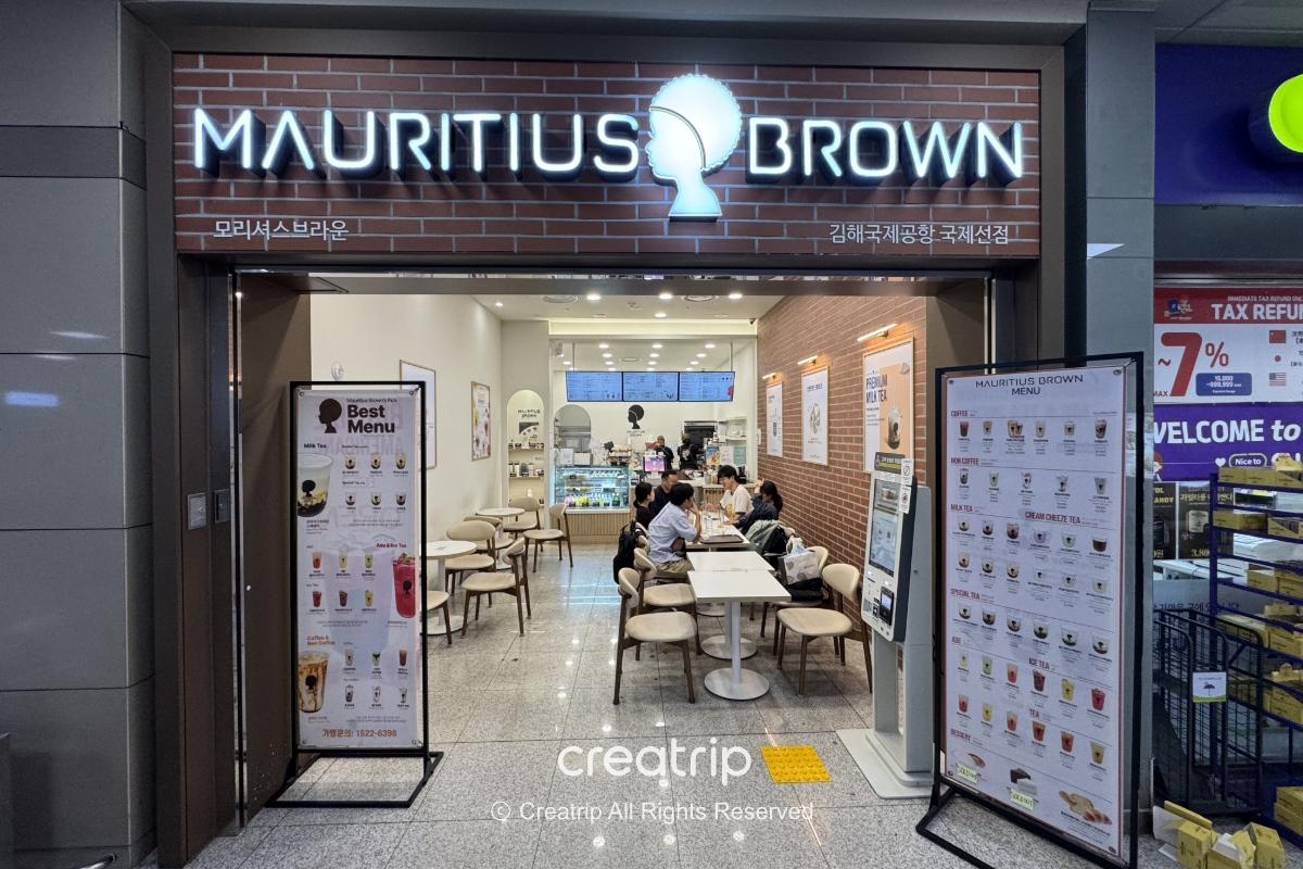 釜山金海机场国际线抵达层的Mauritius Brown饮料店入口与内用座位，招牌奶茶菜单看板、无人点餐机与几位顾客内用情景，适合抵达后买饮品或外带小憩。