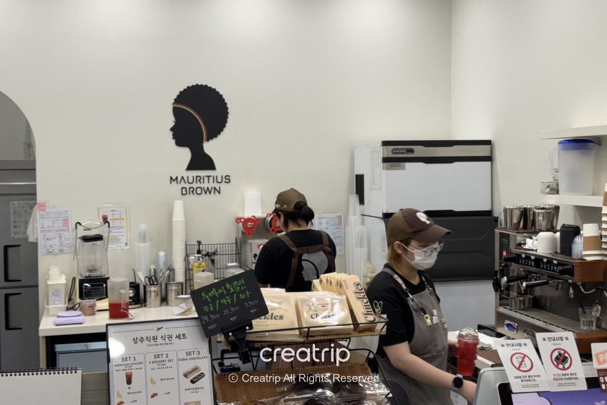 釜山金海机场Mauritius Brown饮品吧台内景，两名穿制服的店员准备奶茶与咖啡，柜台摆放包装点心与自助取餐指示，适合抵达后买饮料或外带轻食。