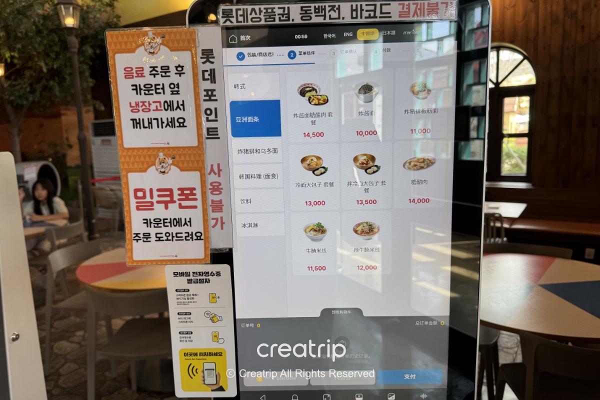 釜山樂天世界美食廣場的自助點餐機畫面，螢幕顯示中英日多語菜單與價位，旁邊有中文與韓文提醒標示，背景可見用餐座位區，突顯點餐便利與家庭用餐氛圍。