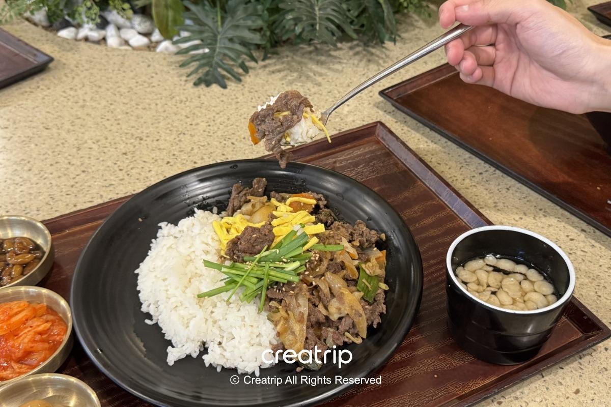 釜山樂天世界Joyful Food Plaza的烤肉蓋飯特寫：白飯與鹹甜烤肉鋪在黑色盤中，旁邊有小菜與清湯，適合家庭遊園時的室內用餐選擇。