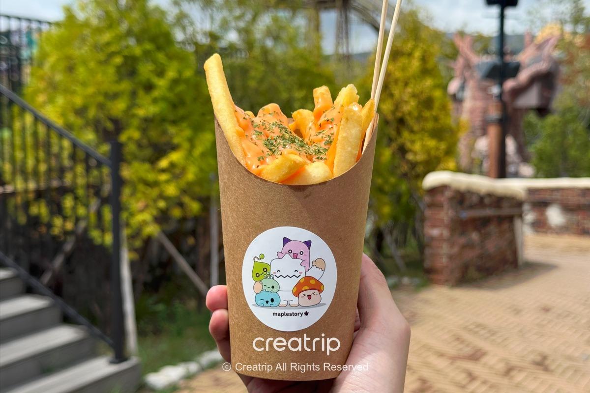 楓之谷聯名起司薯條，紙杯裝上淋滿切達起司與香料的金黃脆薯，背後可見遊樂園步道與綠樹，適合邊拍照邊享用的園區小吃。