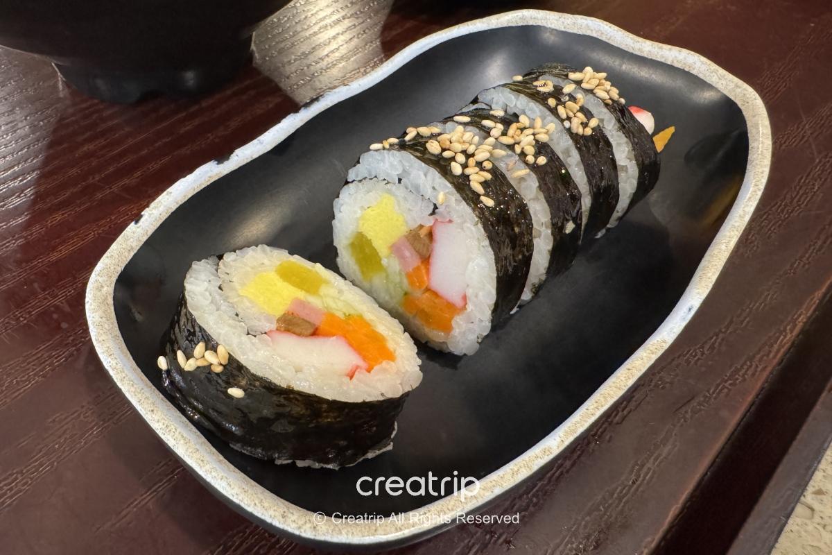 釜山樂天世界Joyful Food Plaza的海苔飯卷切片擺在黑色盤中，內餡有醃黃蘿蔔、蟹肉棒、蛋及蔬菜，表面灑芝麻，適合快速補充能量的園區餐點