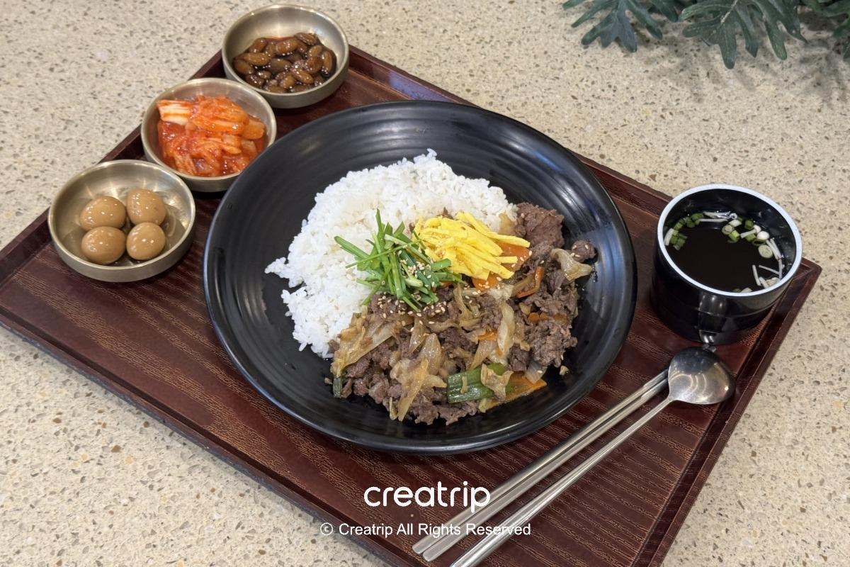 釜山樂天世界Joyful Food Plaza的烤肉蓋飯套餐：黑色碗裝白飯與鹹甜烤肉，旁邊有小菜（泡菜、鵪鶉蛋、醃豆）、清湯與餐具，適合家庭用餐的園區正餐選擇。