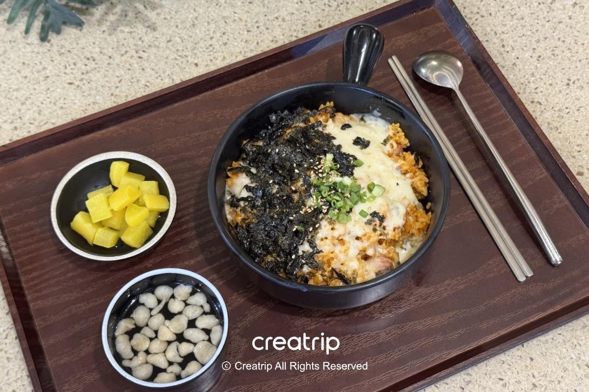 黑色托盤上的起司泡菜炒飯蓋飯，碗內撒有海苔與蔥花，旁邊小碟有黃蘿蔔丁與清湯配餃子，配有韓式筷子與湯匙，呈現樂天世界美食街套餐