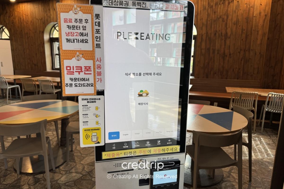 釜山樂天世界Joyful Food Plaza的自助點餐機畫面，中文選單與刷卡裝置可見，背景為寬敞有座位的美食廣場，適合家庭用餐與避暑休息