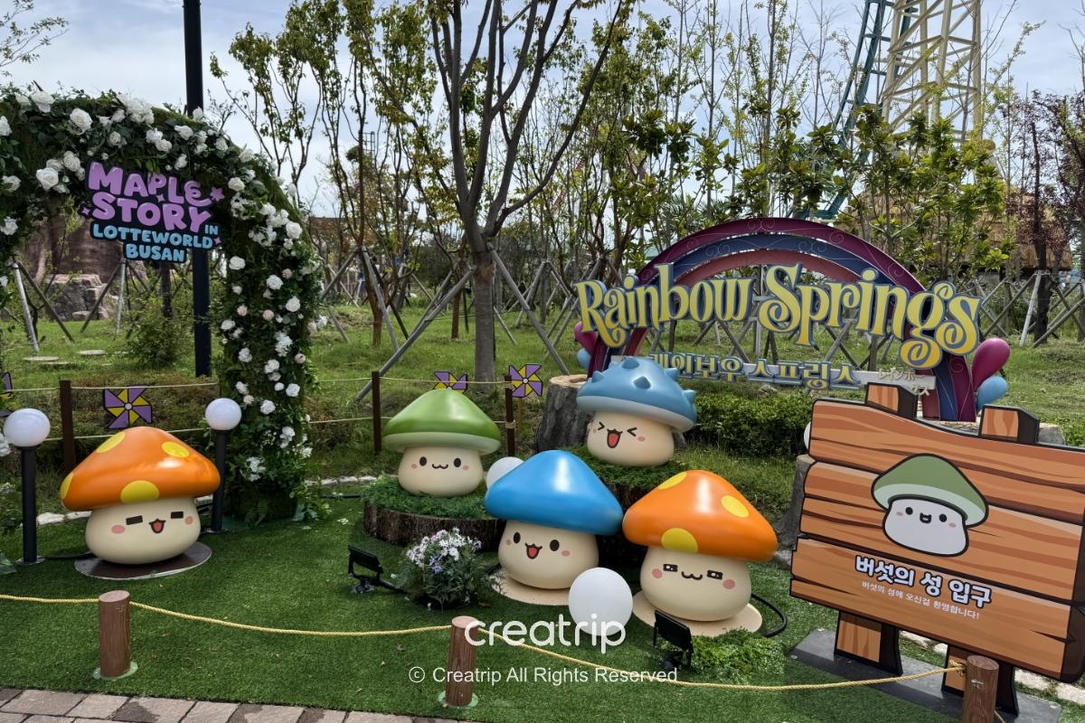 釜山樂天世界楓之谷主題拍照區，草地上擺放多款可愛菇菇造型公仔與Rainbow Springs標誌，適合親子打卡、園區聯名限定小吃導覽圖景