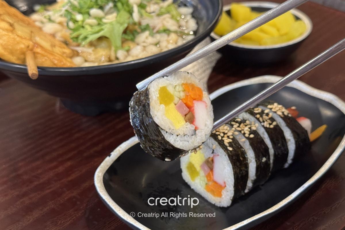 在Joyful Food Plaza點的海苔飯卷切片特寫，筷子夾起一塊可見醃黃蘿蔔、蟹肉棒、胡蘿蔔與米飯，背景有烏龍麵與小菜，呈現遊樂園內快速正餐選擇。