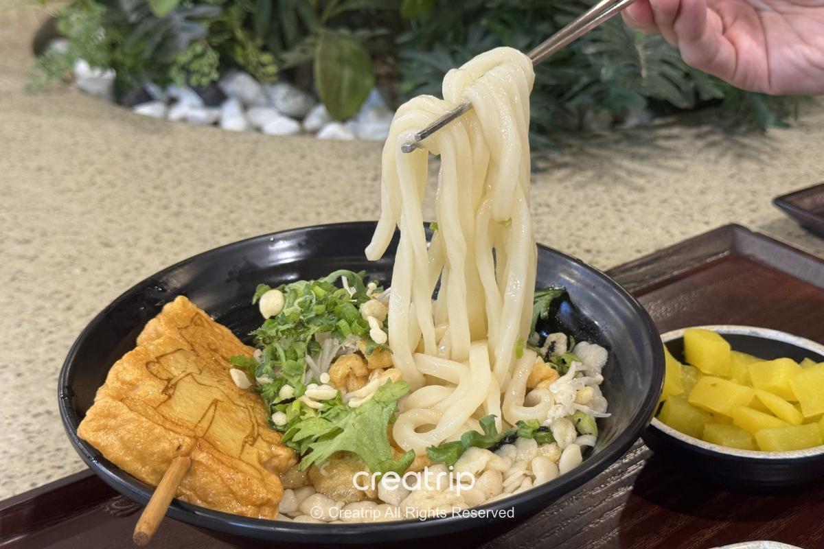 釜山樂天世界Joyful Food Plaza的烏龍麵特寫：筷子夾起彈牙麵條，碗內有魚板、天婦羅碎、蔬菜與小菜白蘿蔔片，適合家庭快速用餐的園區餐點示意