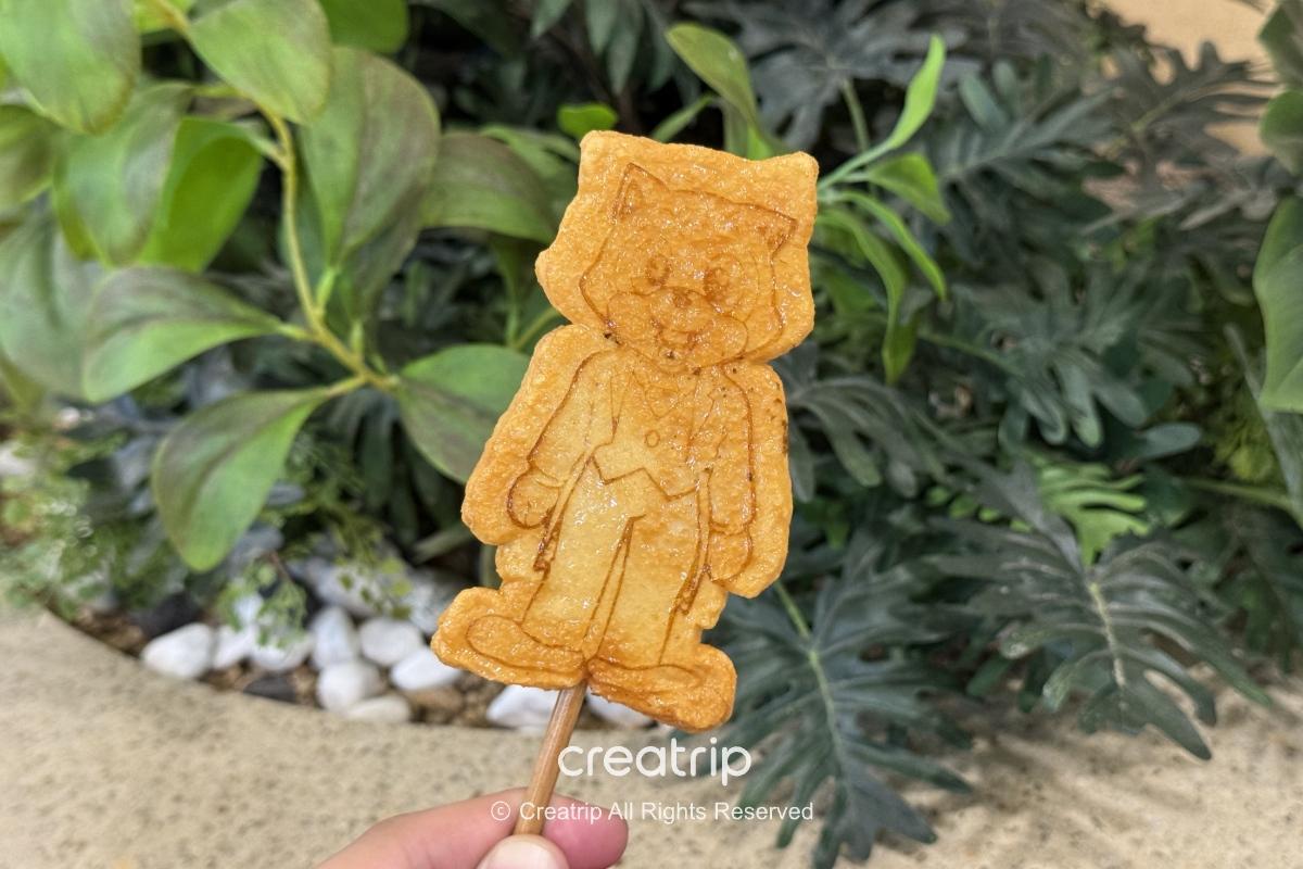 樂天世界造型魚板串，人物造型魚板烤至金黃，插在木棒上拍攝，背景為綠色植物與園區室內地面，適合家庭小朋友點心選擇