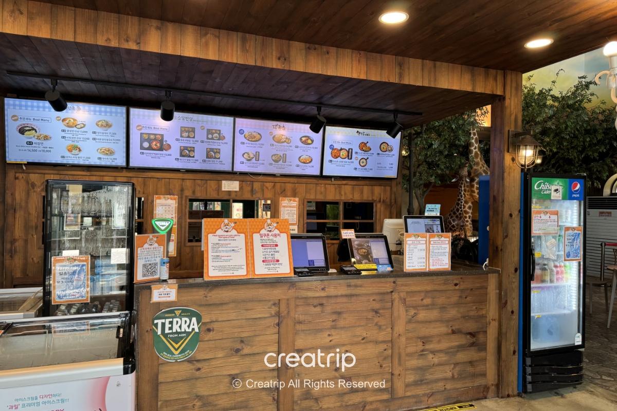 釜山樂天世界Joyful Food Plaza的木質櫃台與自助點餐機，電子菜單牌、冰箱與菜單告示牌一目了然，適合全家坐下用餐的室內美食廣場場景