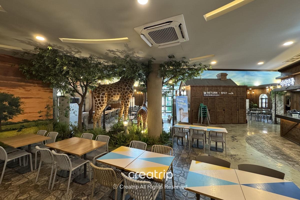 釜山樂天世界Joyful Food Plaza寬敞美食廣場內景，室內座位與桌椅、裝飾性長頸鹿模型與綠植，適合家庭避暑用餐的冷氣座位區畫面