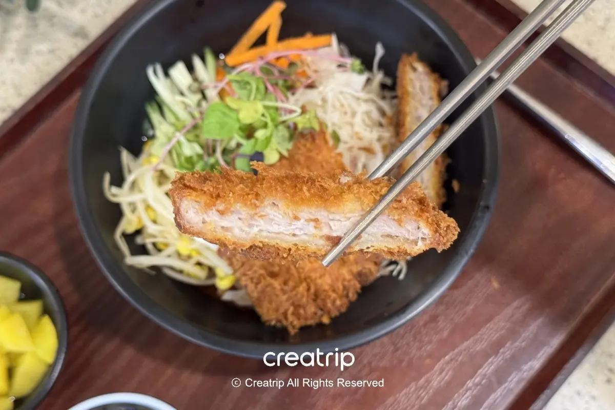 在釜山樂天世界美食廣場的豬排蓋飯特寫，金黃酥脆的炸豬排夾起展現內裡多汁肉質，底下搭配生菜豆芽與飯，旁邊有小菜與湯，適合家庭用餐休息時享用。