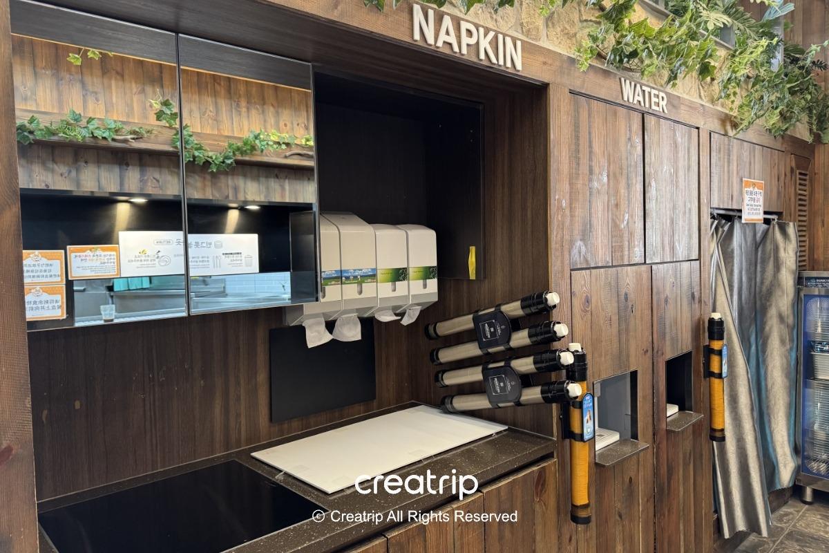 釜山樂天世界Joyful Food Plaza自助取餐區：木質牆面標示NAPKIN與WATER，設有紙巾、飲水取用口以及餐具回收與自助取餐台，適合家庭休息用餐環境。