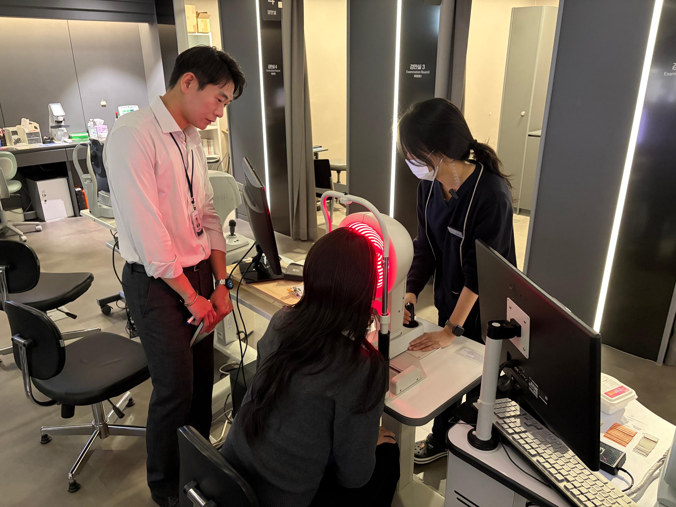 江南眼科檢查中心場景：患者坐在檢查儀前接受角膜或視網膜相關精密檢查，旁有穿制服的醫護與翻譯人員協助說明，環境明亮、儀器排列整齊，突顯當日檢查流程與中文服務陪同