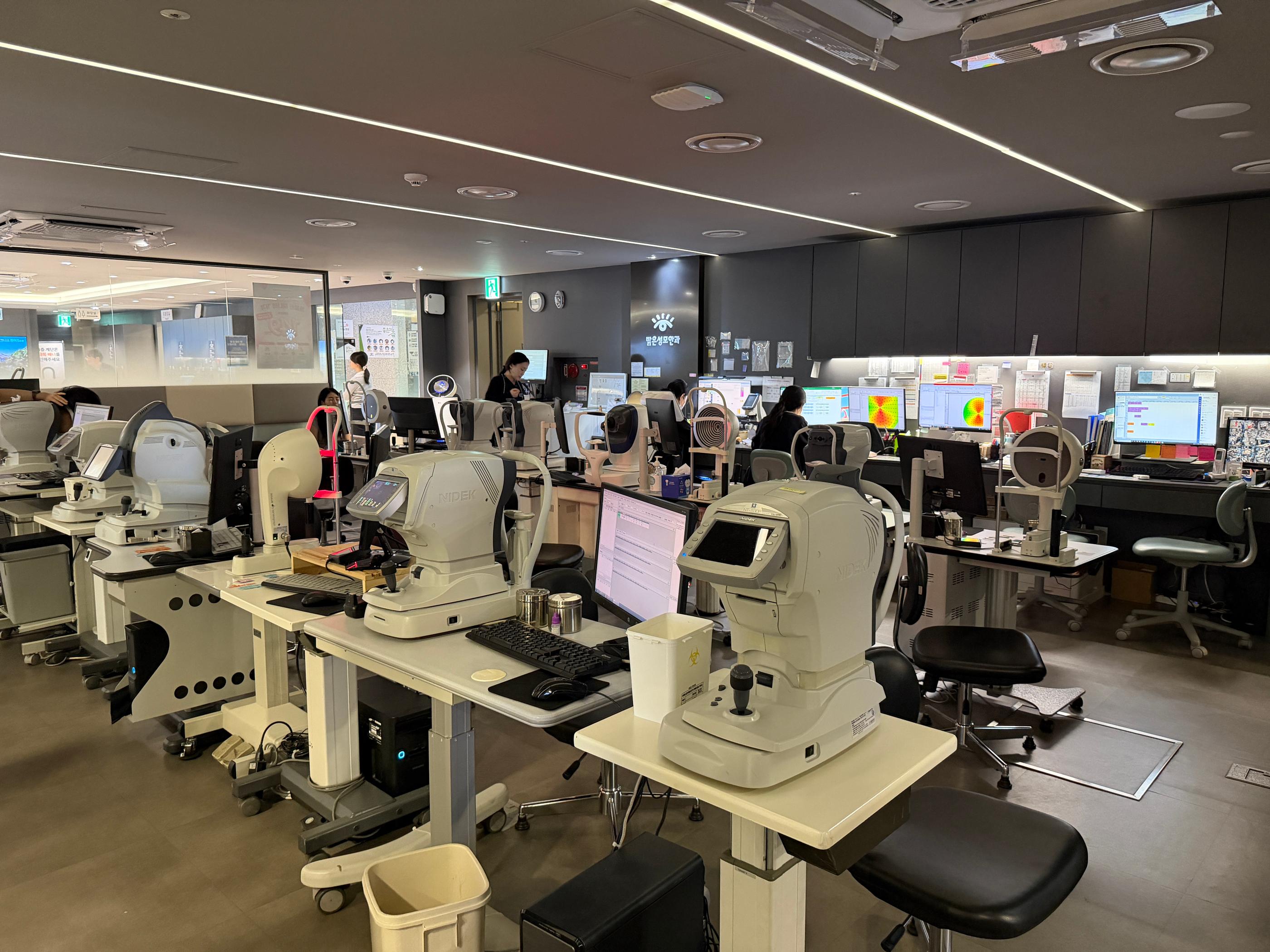 江南明亮聖母眼科的精密檢查中心內景：多排自動化視力與角膜檢測儀器、電腦螢幕與驗光師工作站，呈現系統化術前檢查流程與中文翻譯協助的臨床環境。