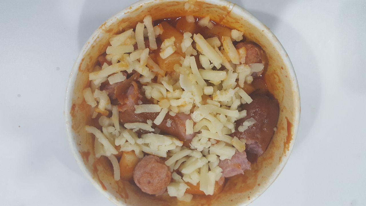 A bowl filled with rice cakes, noodles, sausage, and cheese shreds, highlighting the rich ingredients of Mark meal before heating.