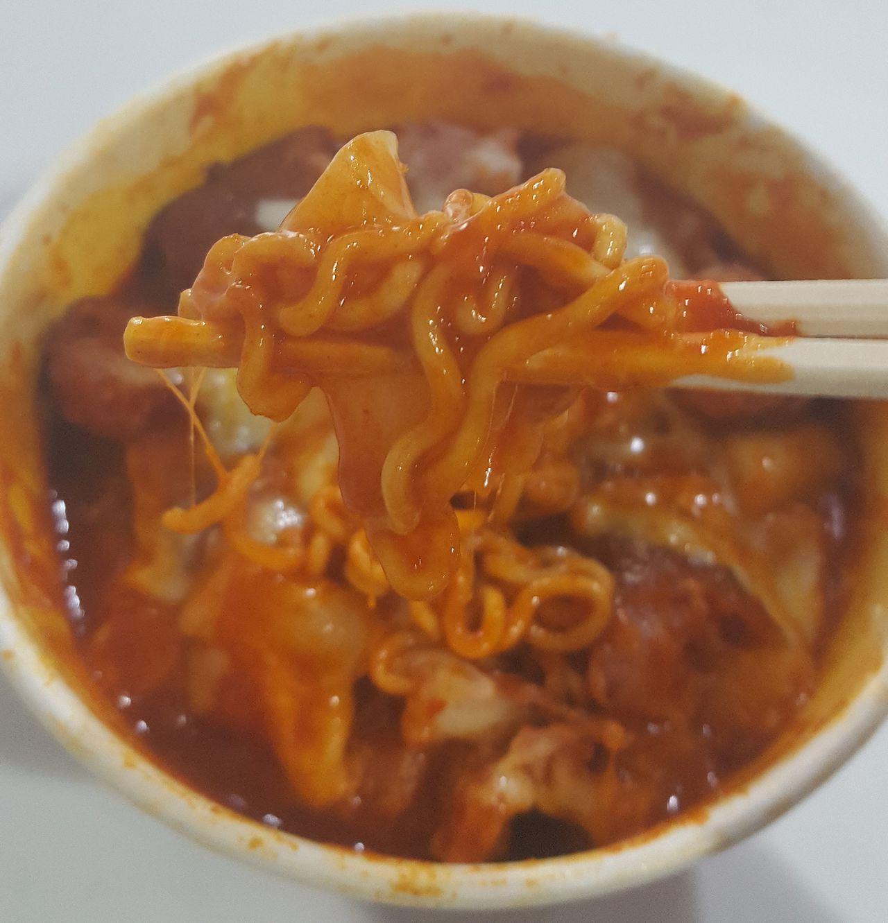 A close-up of a bite of Mark meal being lifted with chopsticks, featuring the cheese-pulled effect and the savory taste of convenience store ingredients.