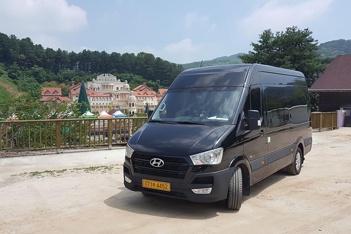 旅行用の大きなバンが屋外の駐車場に停まっている。車は黒色で、韓国の旅客輸送用に一般的に使われるタイプの乗り物です。