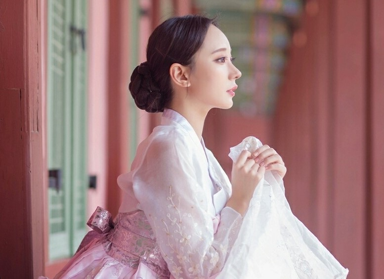 一位女性身穿粉色韓服，坐在染紅色的朝鮮時代建築之中，背景是優雅的木結構。