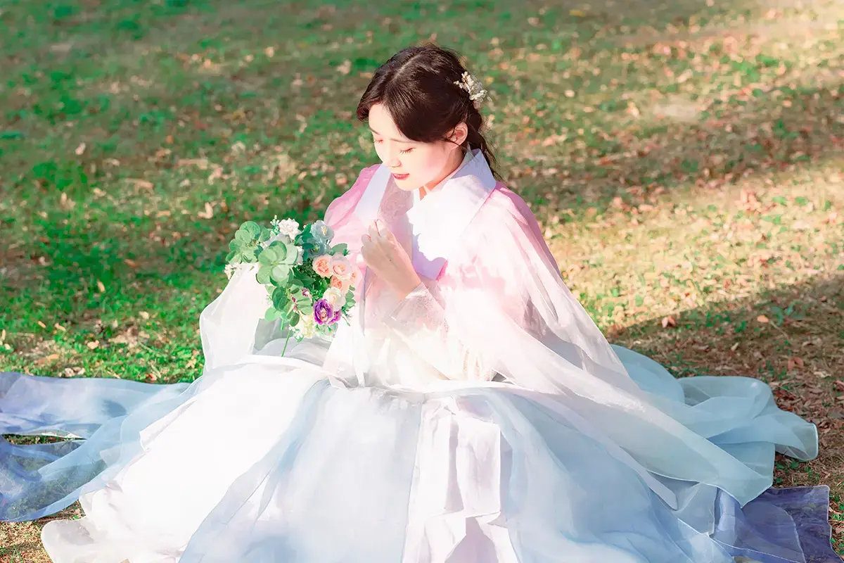 穿著優雅粉色和白色韓服的女性坐在草地上，手捧花束，背景為綠色草坪。