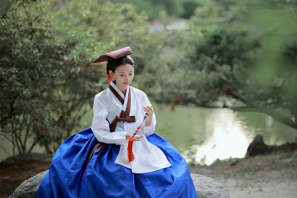 一位年輕女子穿著傳統藍色和白色韓服，坐在石頭上，背景是綠色的園林樹木。