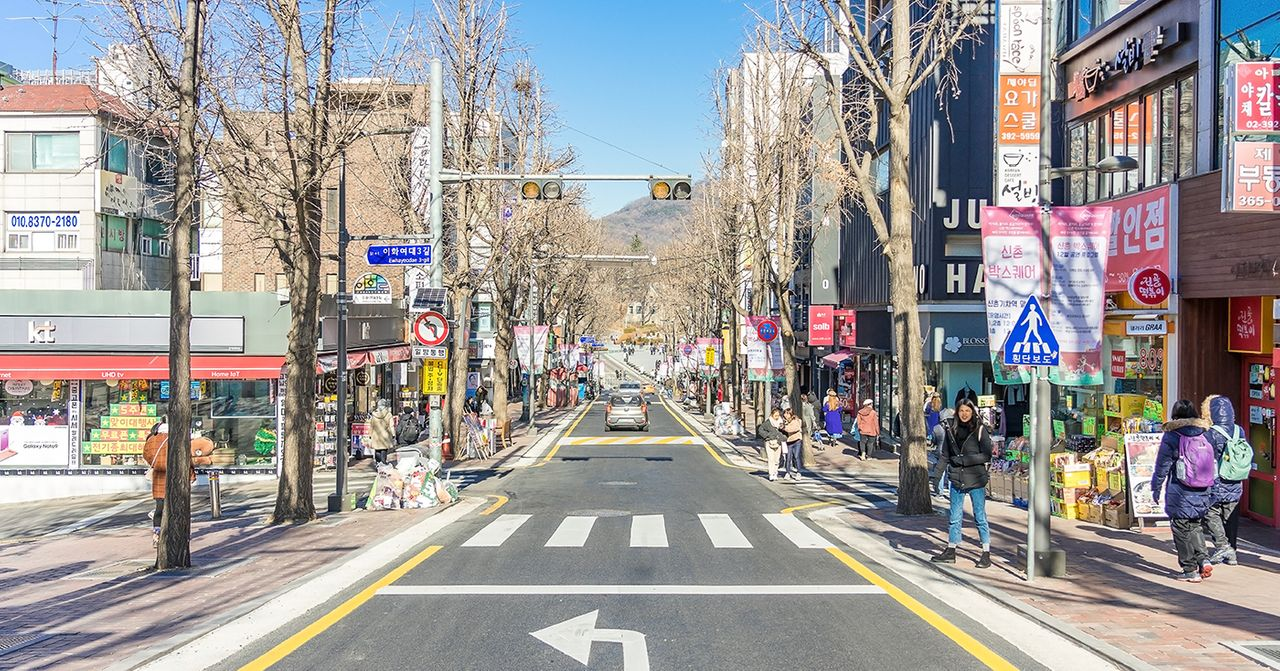 A bustling street in front of Ewha University in Korea filled with shops and pedestrians under a clear blue sky.
