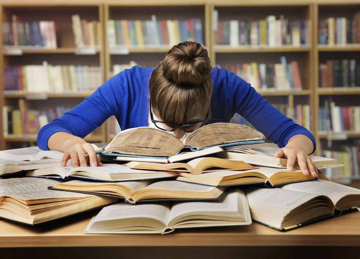 Person overwhelmed with studies, resting on a pile of open books inside a library.