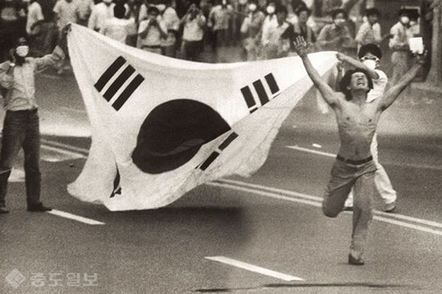 Historic photo of a man running with a large South Korean flag on the street, symbolizing the spirit of protest and resilience during the Gwangju Uprising