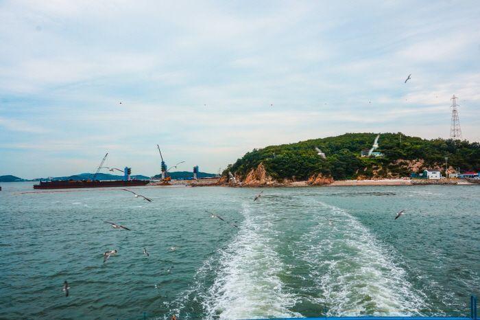 Passengers on a ferry leaving Muido Island, with a scenic view of the island's coastline and seagulls flying overhead.