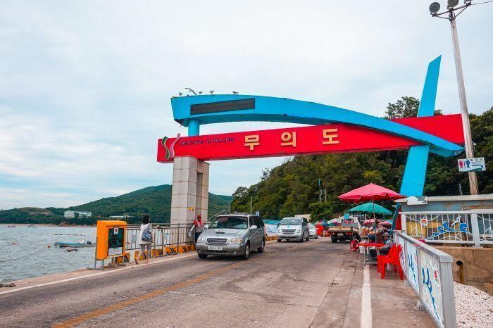 Entrance to Muido Island, marked with a vibrant sign over a road, with nearby stalls and scenic hills in the background.