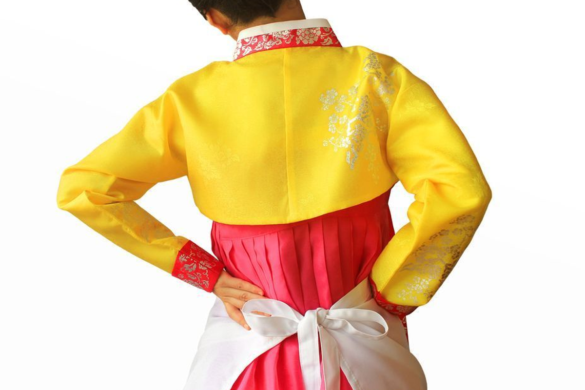 หญิงสวมชุดฮันบกสีเหลืองและชมพู กำลังผูกผ้ากันเปื้อนสีขาวที่เอว แสดงถึงความเป็นงานบ้านที่ไม่มีวันสิ้นสุดในเทศกาลเกาหลี