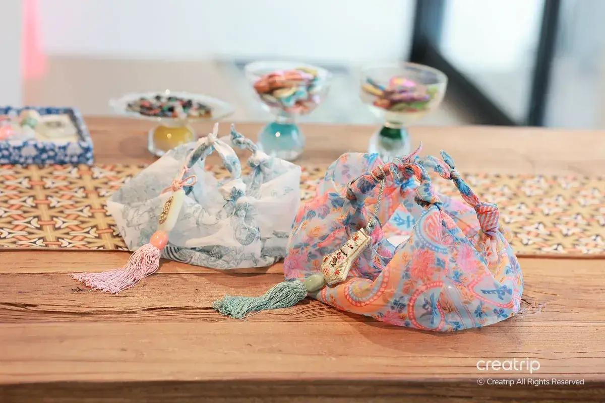 韓式手作體驗作品：兩個彩色綢布束口袋搭配流蘇掛飾，擺放在木桌上，背景模糊可見小碟飾品，適合雨天室內DIY行程示意圖。
