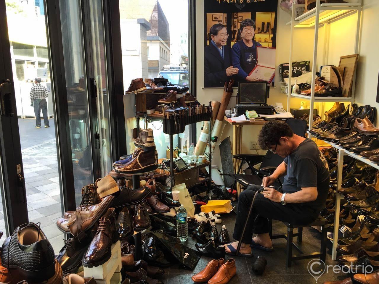 Inside a workshop at the Seongsu-dong handmade shoe street, featuring a skilled artisan crafting leather shoes surrounded by finished shoes on display.