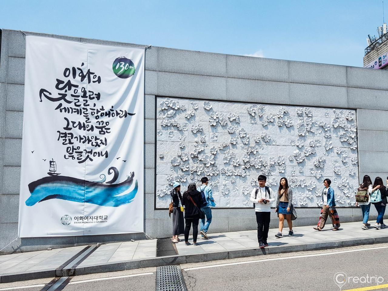 梨花女子大学，墙上有韩国语标语和白色花纹装饰，游客在阳光明媚的日子里行走。韩国首尔著名的大学和观光景点。