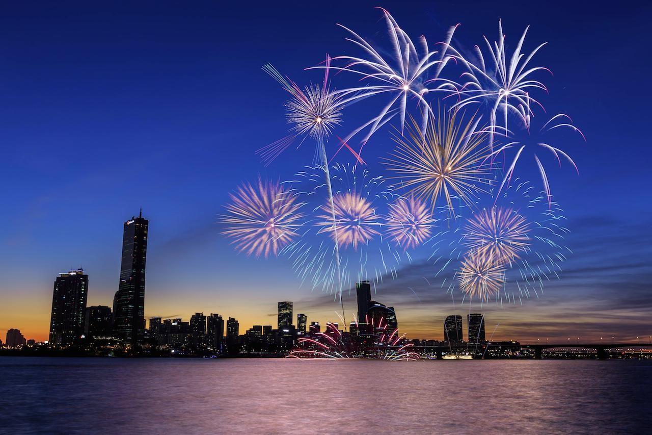 汉江烟火节的夜空场景，烟花在夜晚的首尔天际燃放，江边的摩天建筑物灯火通明，非常适合拍摄的地点。