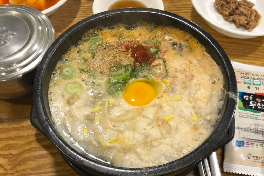 一碗装满豆芽汤饭的砂锅,中间有一个黄色的蛋黄,周围有葱花和其他配料,放在木桌上,旁边有白色的小碟装有小菜。