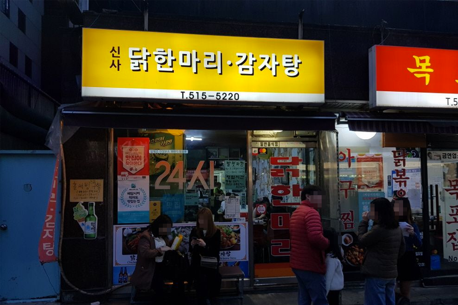一间亮黄色招牌的餐厅外观,上面写着韩文"닭한마리 감자탕"(一只鸡马铃薯汤),门口有几个人在等候,用餐环境显示这是一个24小时营业的地方。