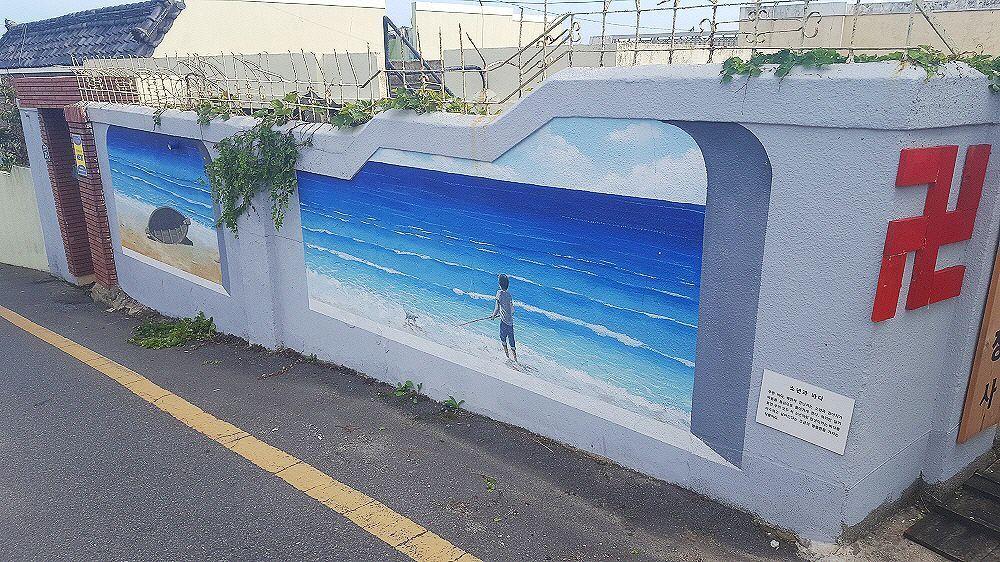 부산 청사포 벽화거리의 벽화 이미지로 파란 하늘과 바다가 그려져 있습니다. 바람이 많이 불고 있는 항구의 장면을 묘사합니다.