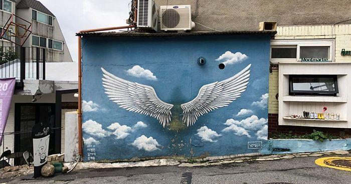 이화 벽화마을의 유명한 천사 날개 벽화 사진. 파란 하늘과 구름이 배경인 푸른 벽에 하얀 천사 날개가 그려져 있습니다.