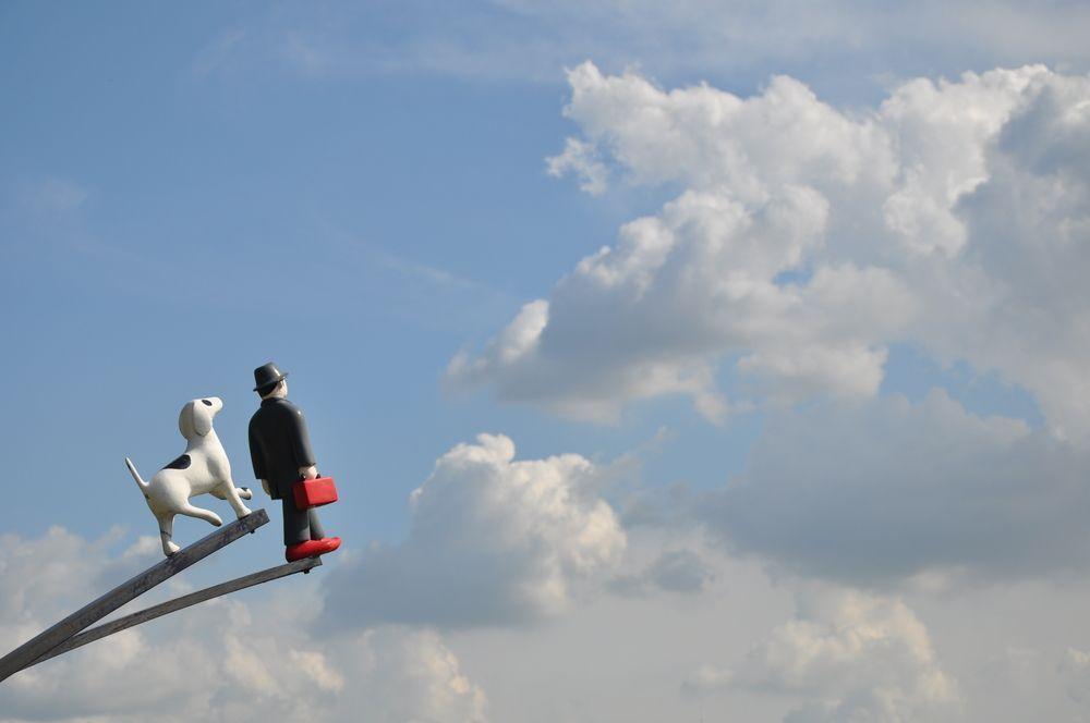 강아지 및 모자를 쓴 남성 조형물이 하늘을 바라보며 펼쳐진 길에 있는 이미지. 화창한 날씨와 하늘이 배경으로 보이는 장면.