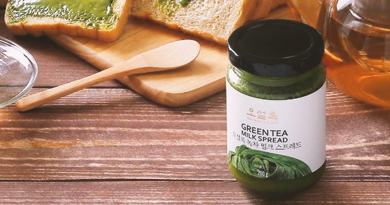 나무 테이블 위에 놓여 있는 녹차 스프레드와 녹차 스프레드를 바른 식빵 조각의 클로즈업 이미지입니다. 투명한 차 잔의 일부분도 살짝 보입니다.