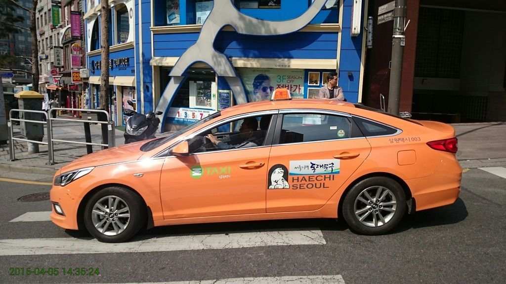 韓國首爾的士圖片，橙色的士車身上寫有“Haechi Seoul”字樣，旁邊是藍色建築物。