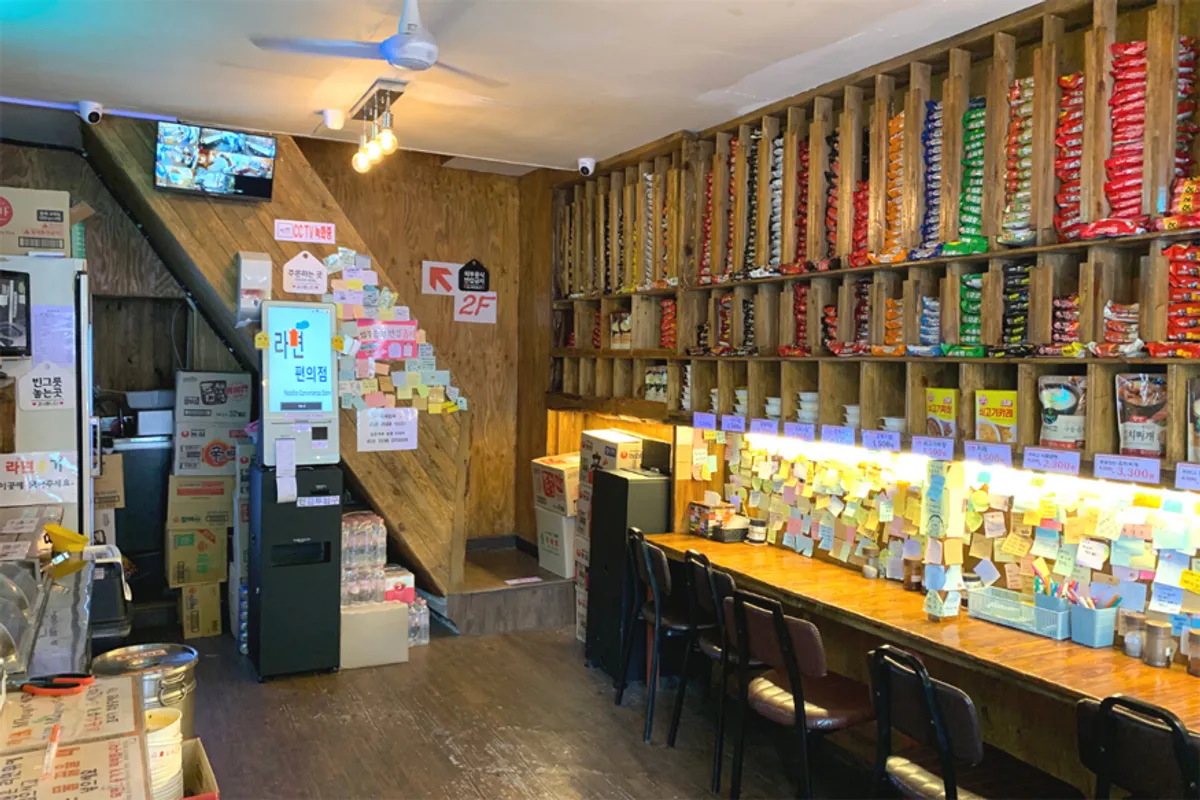 Interior of a Korean restaurant with self-service concept, featuring rows of instant noodle packages on display and tables for single diners.