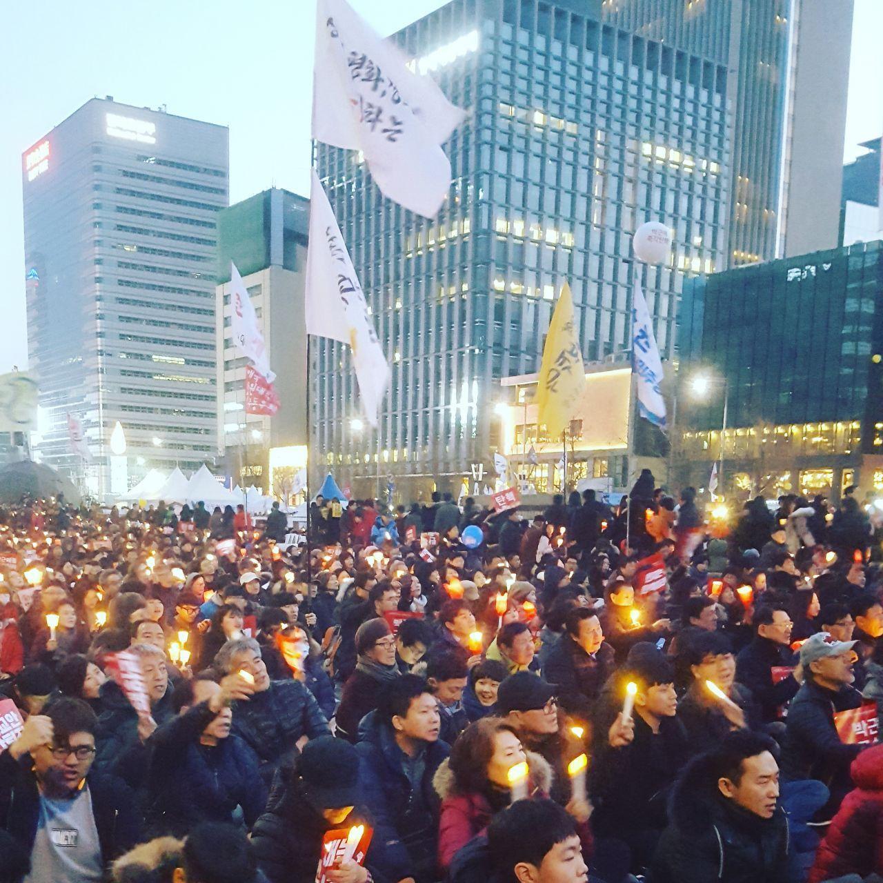 Photo of a packed protest in the city, with people holding candles and banners, symbolizing civic activism and discontent in Korea.