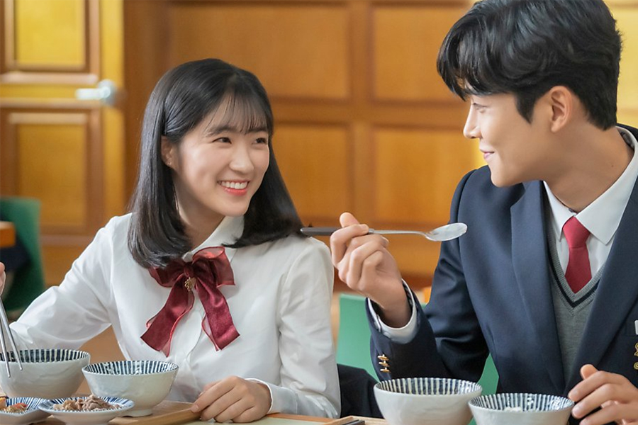 Two Korean high school students in winter uniforms sitting at a table during lunchtime, showcasing traditional Korean school attire with a focus on their formal wear and dining setting.