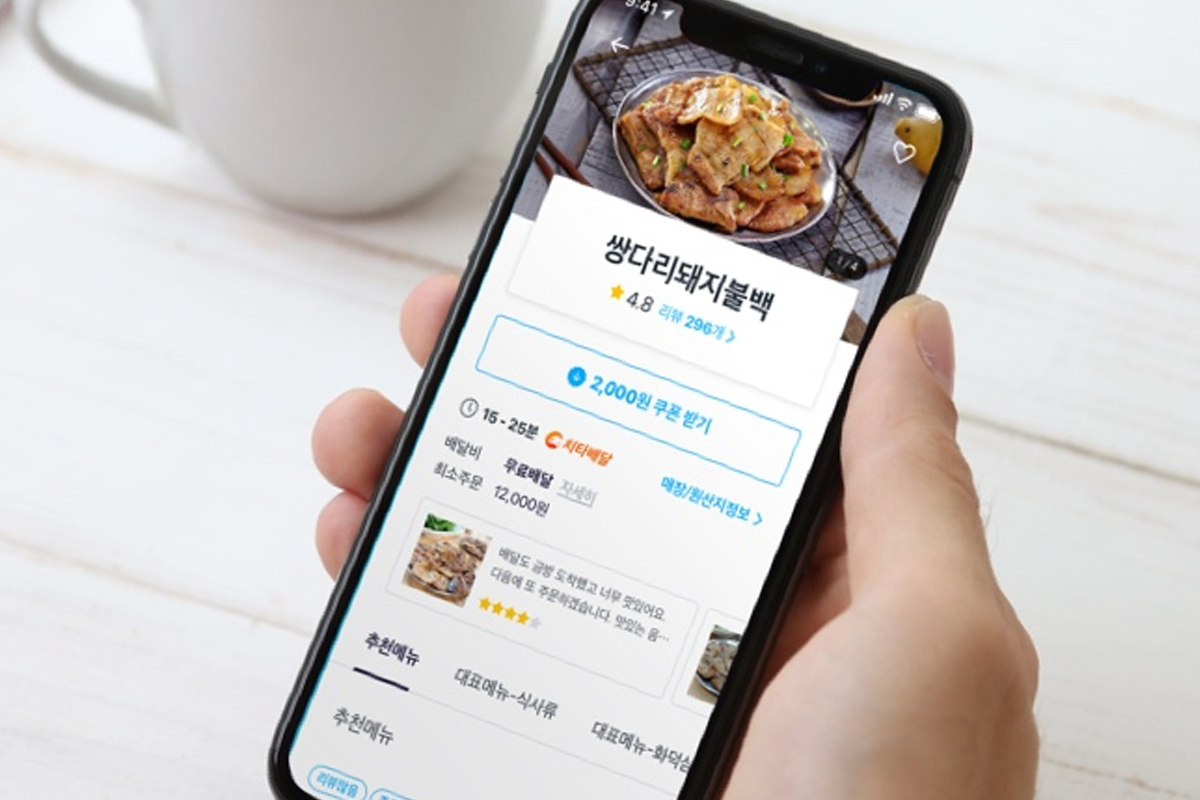 手持手機顯示韓國外送App餐點頁面，畫面包含菜名、評分、價格與到貨時間，搭配桌上咖啡杯，突顯旅客在韓國使用外送服務的情境與操作介面。